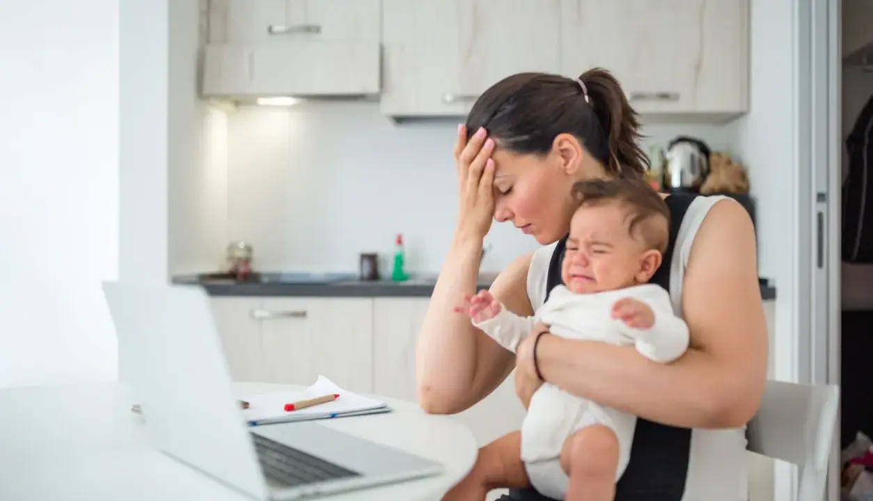 Zmęczona mama z płaczącym niemowlakiem na rękach, zastanawia się, przebodźcowanie niemowlaka ile trwa. Obok laptop i dokumenty.