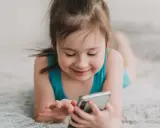 Uśmiechnięta dziewczynka leży na dywanie, bawiąc się telefonem. Wpływ telefonu komórkowego na mózg dziecka jest tematem dyskusji.