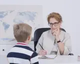 Chłopiec rozmawia z nauczycielką o ocenach. Nauczycielka słucha uważnie, notując coś na kartce.