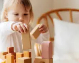 Mała dziewczynka, która ma 4 latka, skupiona buduje wieżę z drewnianych klocków, nie zwracając uwagi na inne dzieci.