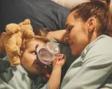 Mama opiekuje się dzieckiem z rotawirusem. Podaje mu wodę z kubeczka, a obok leży pluszowy miś.