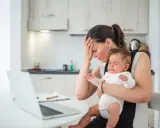 Zmęczona mama z płaczącym niemowlakiem na rękach, zastanawia się, przebodźcowanie niemowlaka ile trwa. Obok laptop i dokumenty.