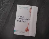 Książka "Niska samoocena u dzieci" Margot Sunderland. Na okładce fragment twarzy dziecka ukrywającego się za białą zasłoną.