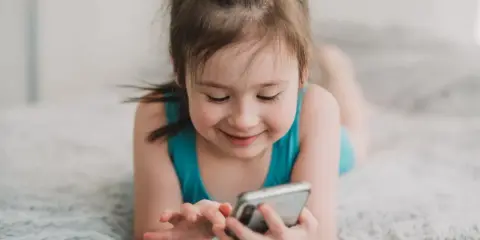 Uśmiechnięta dziewczynka leży na dywanie, bawiąc się telefonem. Wpływ telefonu komórkowego na mózg dziecka jest tematem dyskusji.