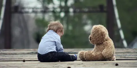 Chłopiec siedzi na drewnianej kładce z pluszowym misiem. Czy dziecko pamięta traumę?