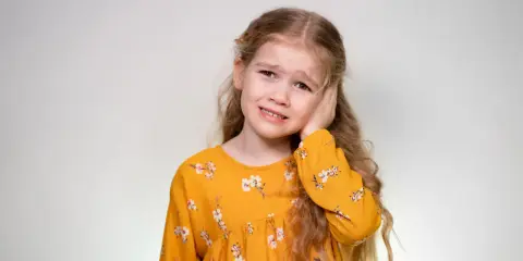 Mała dziewczynka w żółtej sukience z kwiatami trzyma się za ucho, jakby moje dziecko wydaje dziwne dźwięki i ją to niepokoi.