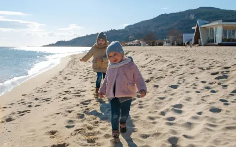 Dwa uśmiechnięte dzieci biegają po piaszczystej plaży. Słońce odbija się od wody, a w tle widać wzgórza i budynek.