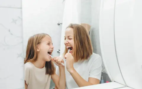 Mama i córka myją zęby, śmiejąc się. Dziecko nie chce myć zębów, ale mama przekonuje je zabawą.