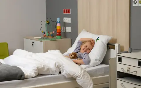 Uśmiechnięte dziecko w szpitalnym łóżku, przytulające misia. Obok zabawka edukacyjna i szafka.