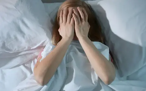 Nastolatka w łóżku, zakrywająca twarz dłońmi, symbolizuje bezsenność u nastolatków i jej przyczyny.