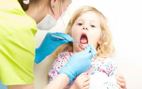 Mała dziewczynka z otwartymi ustami podczas pierwszej wizyty u dentysty. Dentystka w niebieskich rękawiczkach bada jej ząbki.
