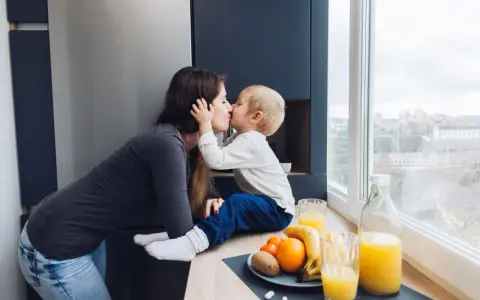 Matka całuje dziecko, siedząc przy oknie. Na blacie owoce i soki.