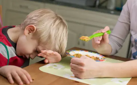 Dziecko z brakiem apetytu, sennością i zmęczeniem odwraca głowę od jedzenia. Mama próbuje nakarmić go łyżeczką.