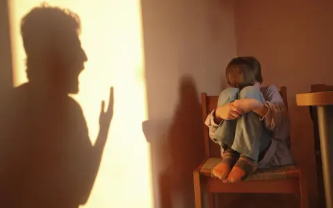 Cień krzyczącej postaci rzuca się na ścianę, podczas gdy dziecko skulone na krześle odczuwa skutki bicia dzieci w dorosłym życiu.