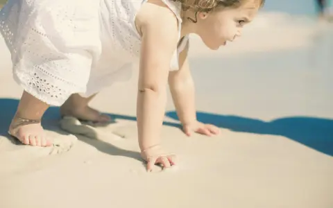 Małe dziecko w białej sukience raczkuje po plaży. Może to być jedna z przyczyn nie chodzenia dziecka, bo piasek jest ciekawy.