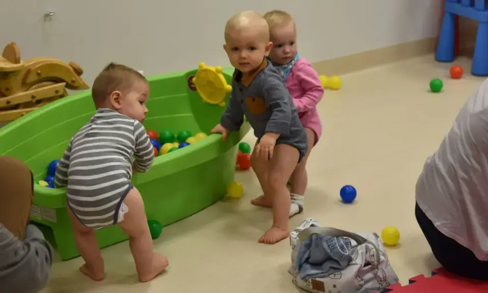 Trzy maluchy, w tym 6 miesięczne dziecko w chodziku, bawią się kolorowymi piłeczkami w zielonej wanience.