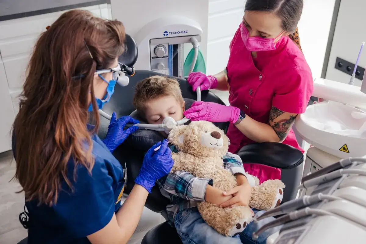 Dziecko śpi spokojnie podczas leczenia zęb&oacute;w pod narkozą, przytulając misia. Dentystki dbają o jego komfort i bezpieczeństwo.