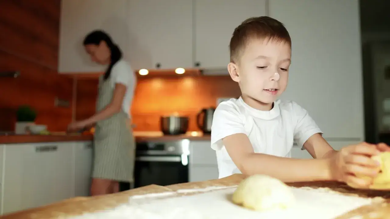 Chłopiec z mąką na nosie lepi ciasto. Mama w tle gotuje. Tak można zachęcić 10 letnie dziecko do nauki przez zabawę.