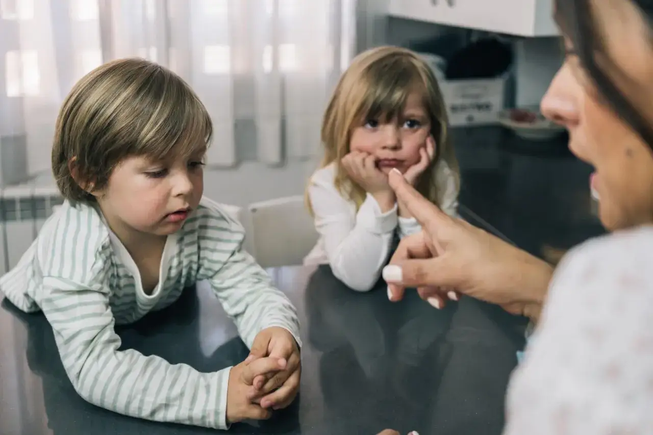 Mama karci dziewczynkę, która opiera głowę na dłoniach, podczas gdy chłopiec patrzy w dół. Wygląda, jakby rodzice pomagali tylko jednemu dziecku.