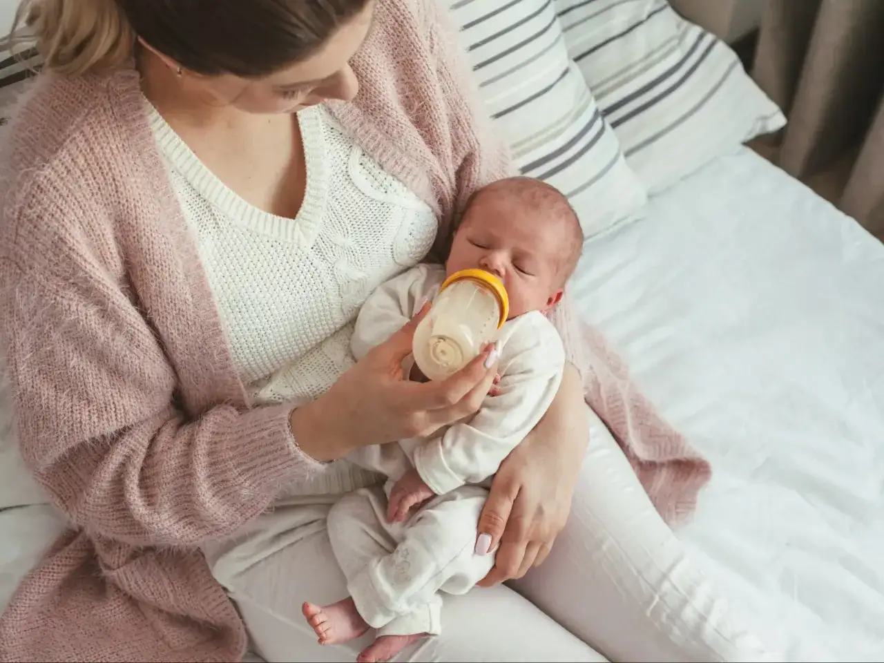 Mama karmi butelką śpiące dziecko. Maluch lekko szarpie się przy karmieniu, ale mama cierpliwie go uspokaja.